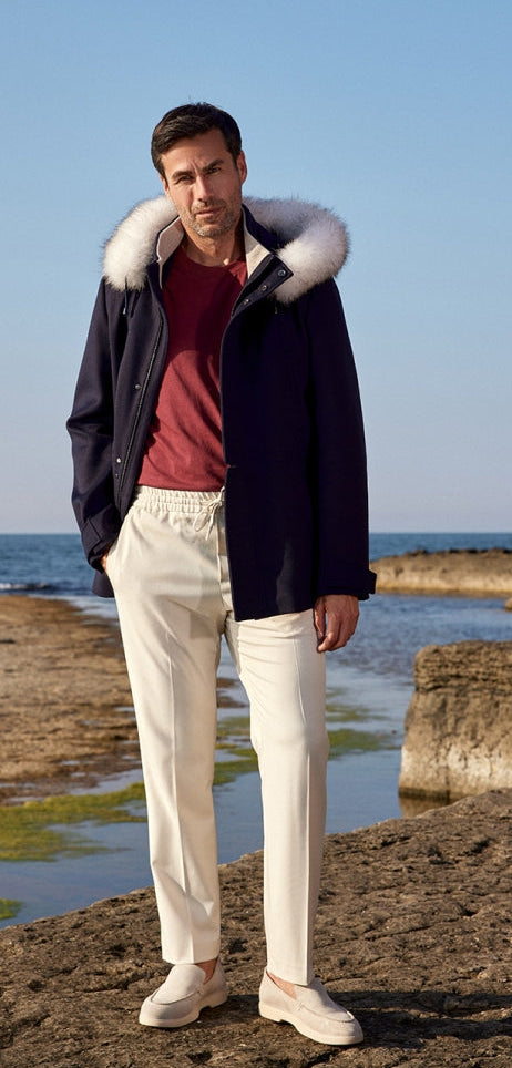 Man standing on a rocky coastal area wearing a dark jacket with a fur-lined hood, red shirt, and white pants.