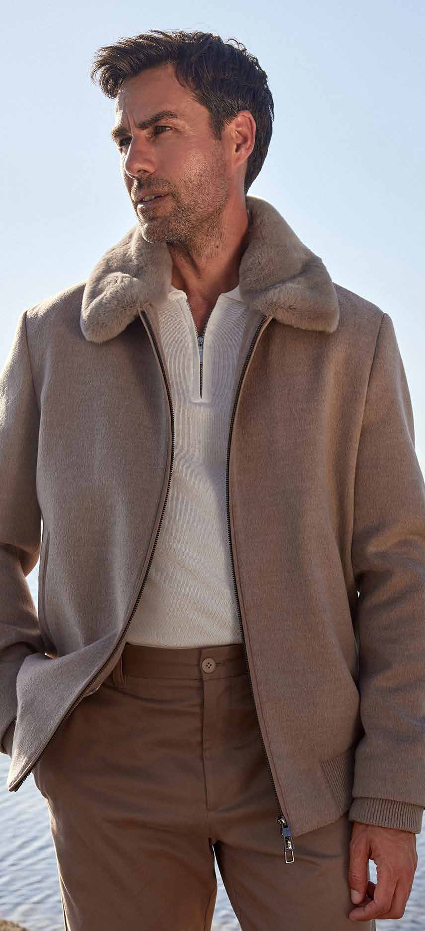 Man wearing a beige coat with a fur collar and brown pants against a clear sky.