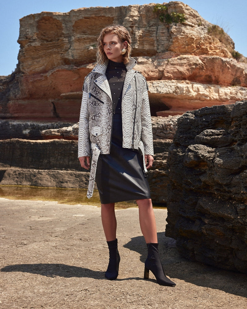 Woman standing in front of rock formations wearing a patterned coat and black skirt.