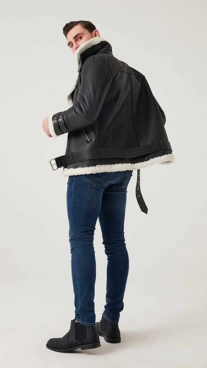 Man wearing a dark jacket with white fur lining, blue jeans, and black boots on a light gray background