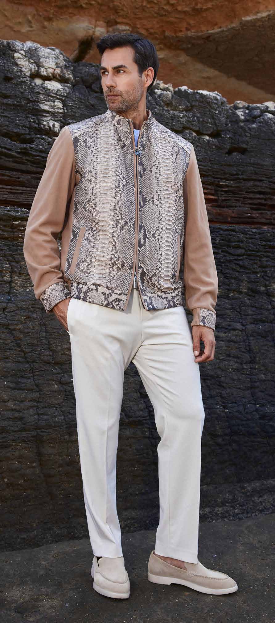Man wearing a patterned vest over a beige shirt with white pants against a rocky background
