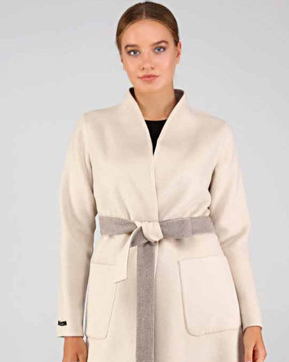 Woman wearing a long cream alpaca overcoat with a belt on a plain background