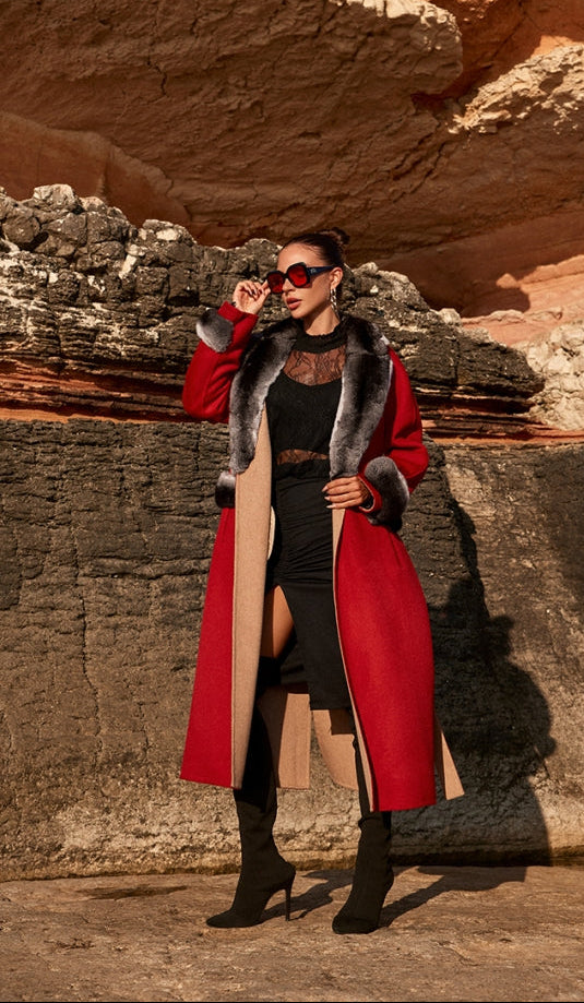Woman wearing a red coat with fur trim standing in front of a rocky wall.