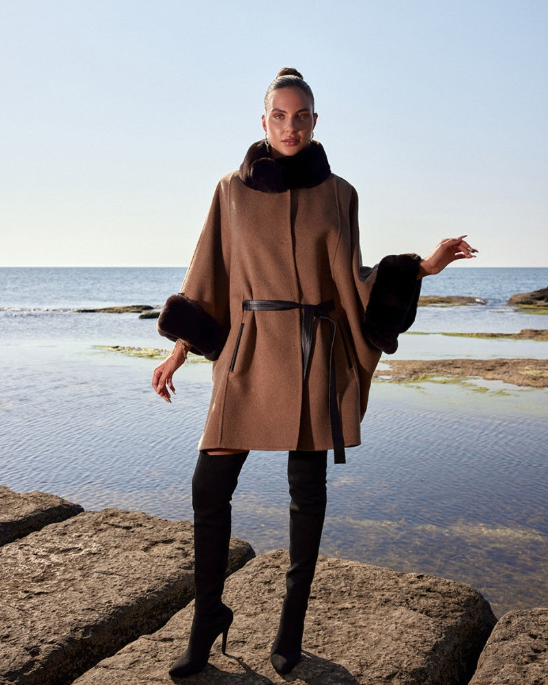 Woman wearing a brown coat with fur trim standing on rocks by the water.
