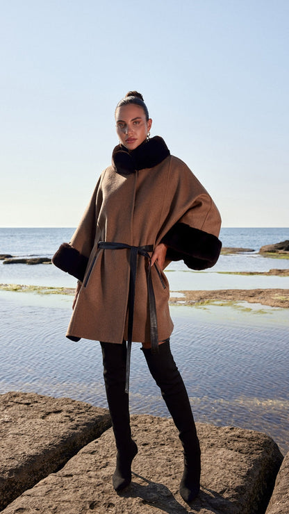 Woman wearing a brown coat with black fur trim standing on rocks by the water.