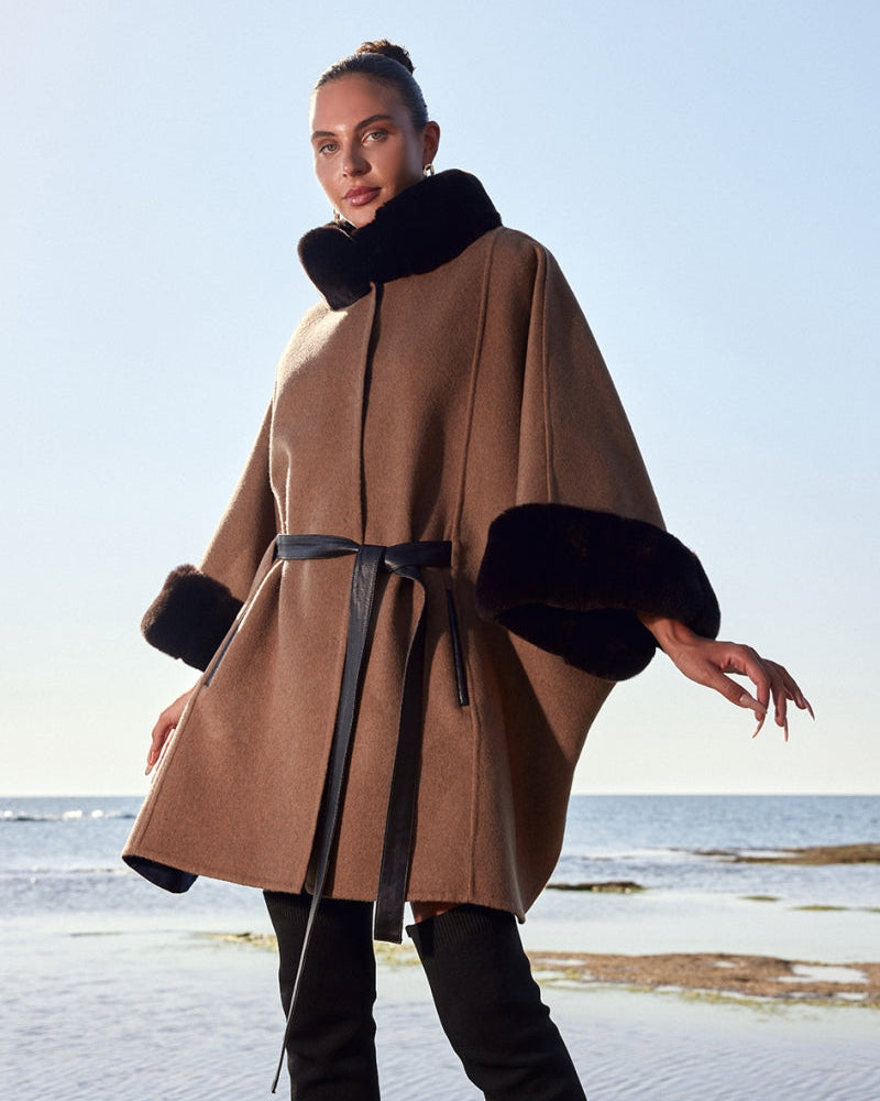 Woman wearing a brown coat with black fur trim standing by the ocean.