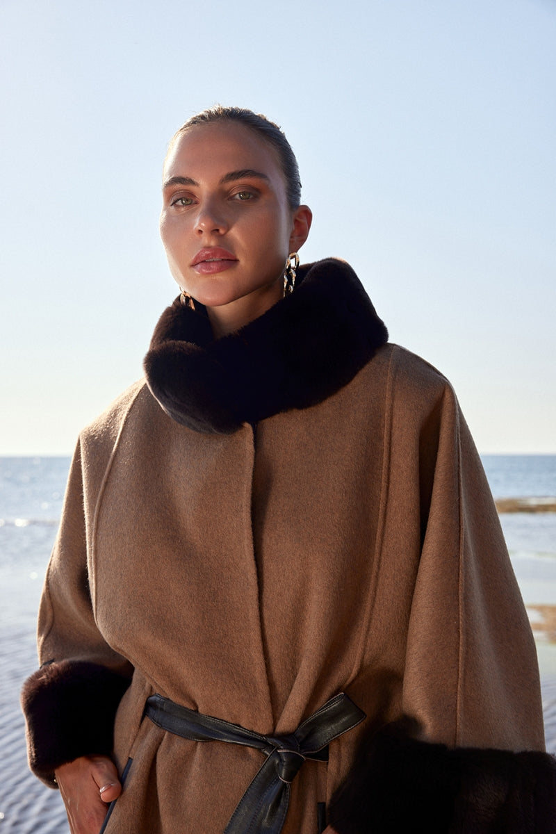 Woman wearing a brown coat with fur trim standing by the ocean.