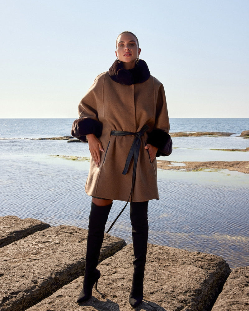 Woman wearing a beige coat with fur trim standing on rocks by the water.