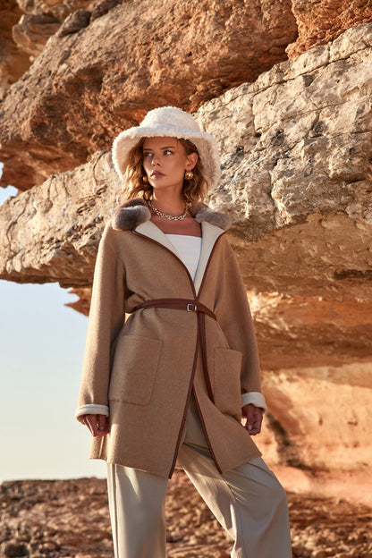 Woman in a beige coat and white hat standing in front of rocky cliffs.