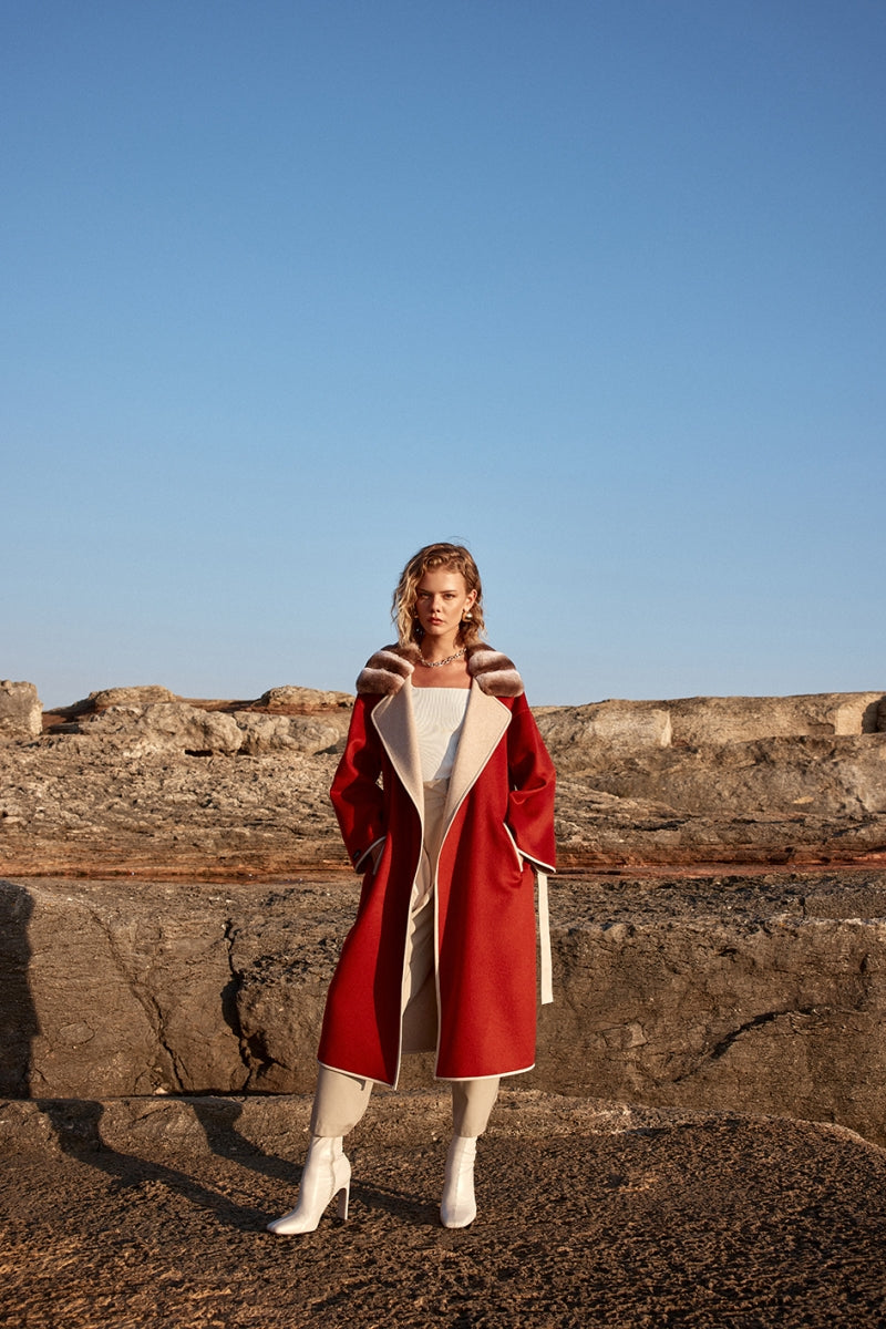 Woman wearing a red coat and white shoes standing on rocky terrain with a clear blue sky.