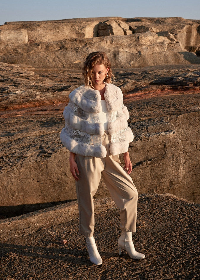 Woman wearing a white fur coat and beige pants standing on rocky terrain.