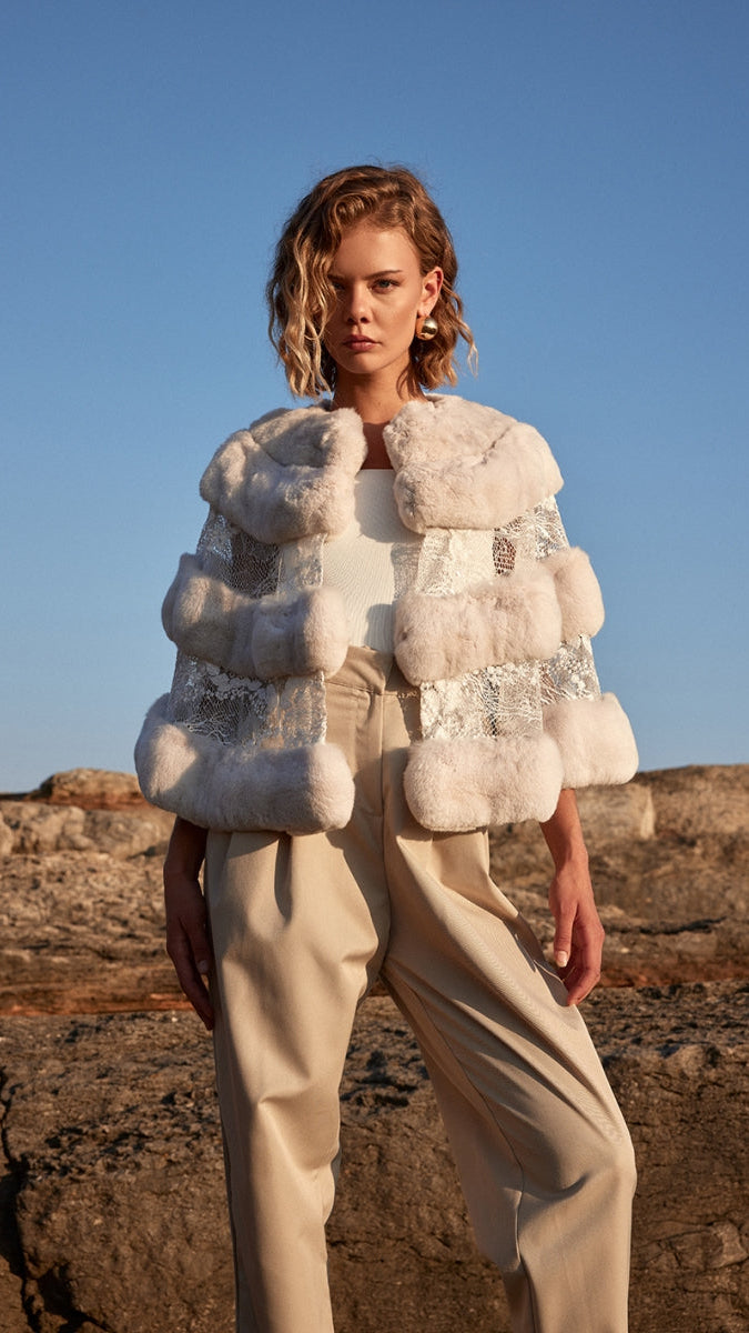Woman wearing a fur vest and beige pants standing against a clear blue sky.
