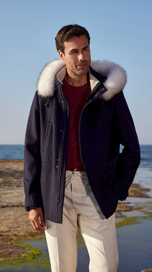 Man wearing a dark coat with a fur-lined hood standing on a rocky shoreline.