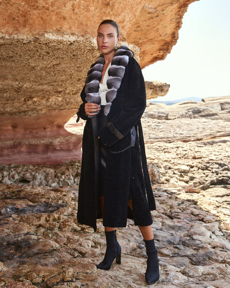 Woman in a long black coat with fur collar standing in a rocky landscape.