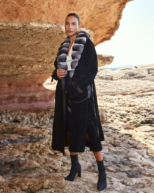 Woman in a long black coat with fur collar standing in a rocky landscape.