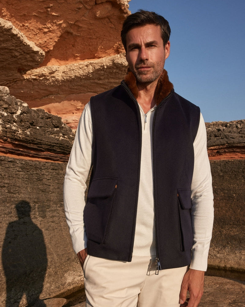 Man wearing a navy vest over a white shirt with beige pants, standing against a rocky cliff background.