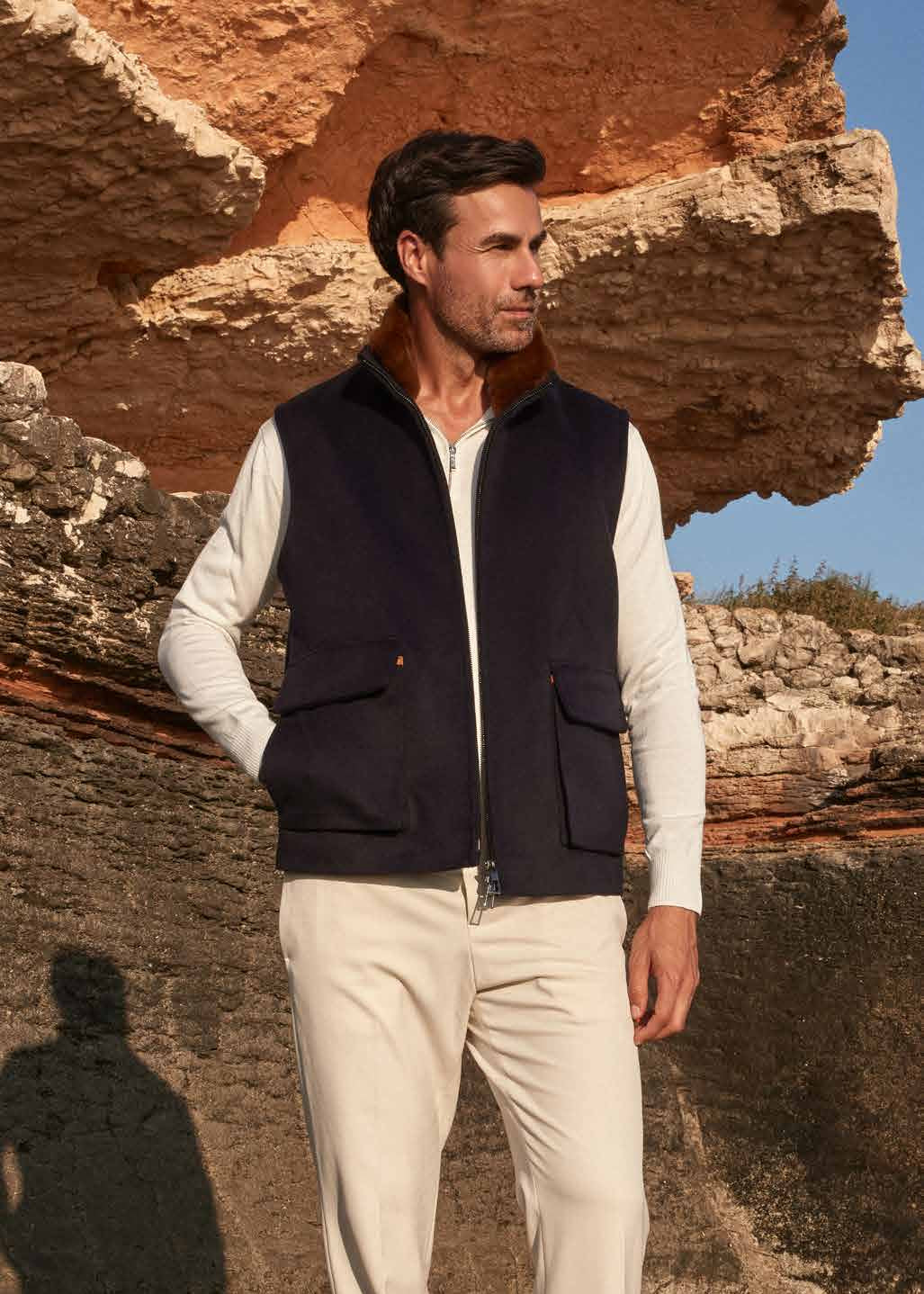 Man wearing a dark vest over a light shirt with beige pants, standing in front of a stone wall.