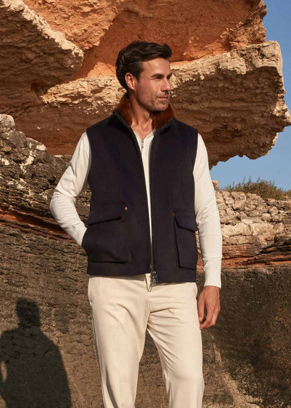 Man wearing a dark vest over a light shirt with beige pants, standing in front of a stone wall.