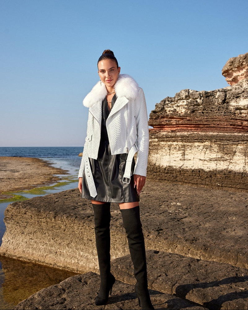 Woman in a white coat and black leather skirt standing on rocky cliffs by the sea.