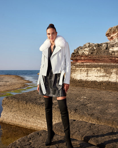 Woman in a white coat and black leather skirt standing on rocky cliffs by the sea.