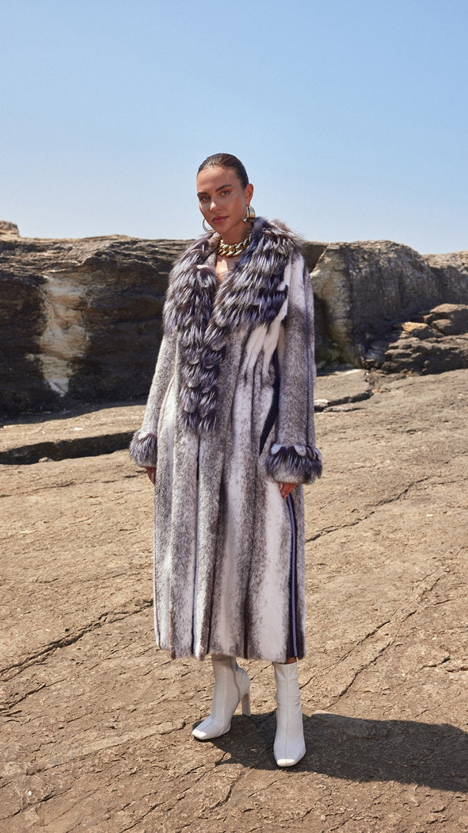 Woman wearing a fur coat and striped dress standing on rocky terrain with clear blue sky.