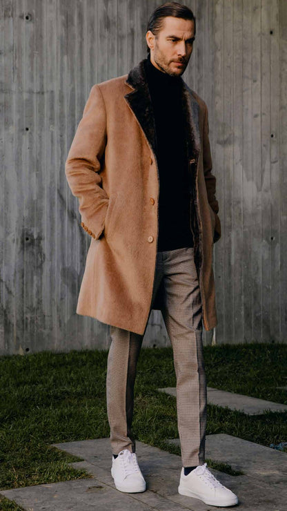 Man wearing a brown coat, black turtleneck, gray pants, and white sneakers against a concrete wall.