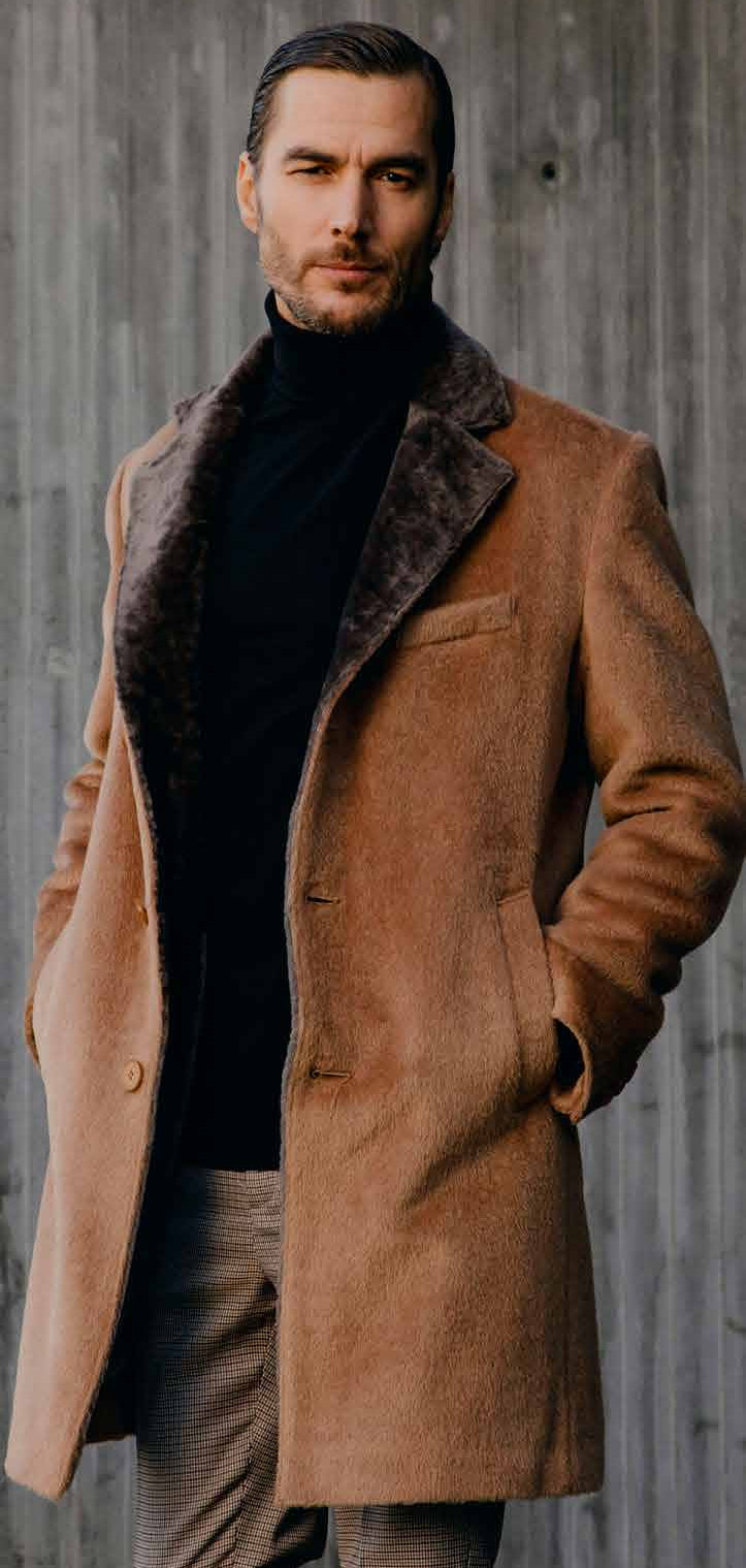 Man wearing a brown coat with fur collar against a concrete wall.