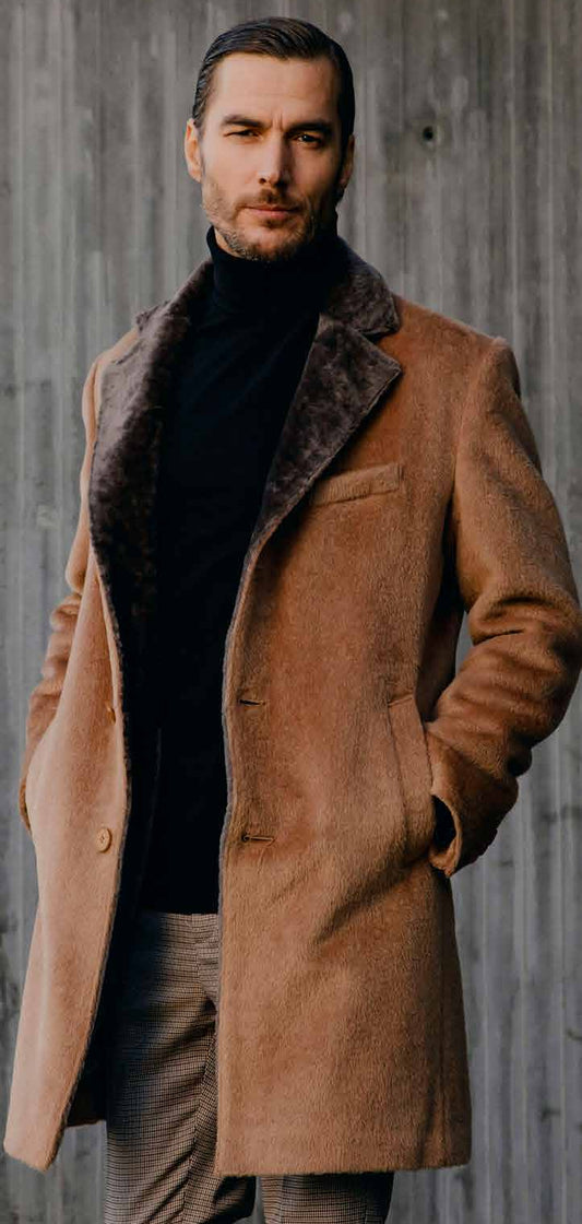 Man wearing a brown coat with fur collar against a concrete wall.