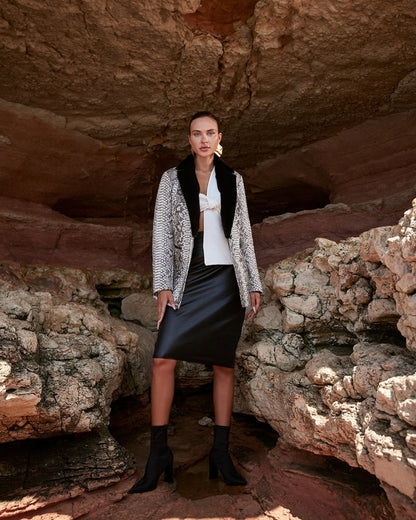 Woman standing in a rocky cave wearing a patterned jacket, black skirt, and white top.