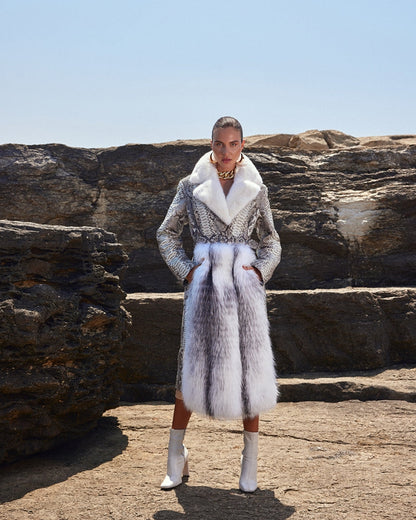 Woman wearing a fur coat and white boots standing on rocky terrain with clear blue sky.