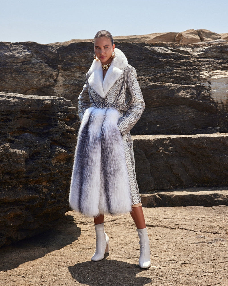 Woman wearing a stylish outfit with a fur coat and patterned dress against a rocky background.