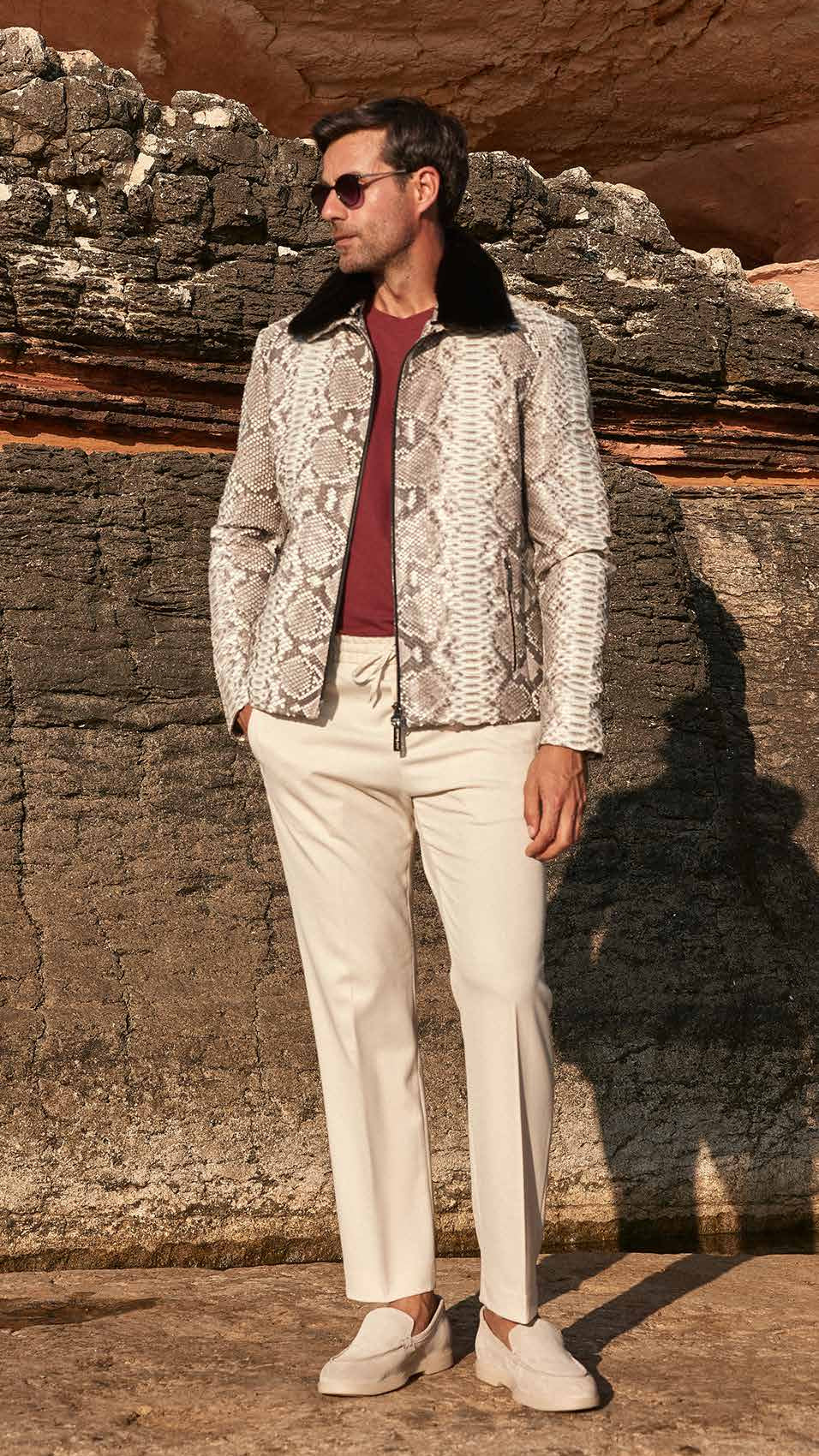 Man standing against a rocky wall wearing a patterned jacket and white pants.