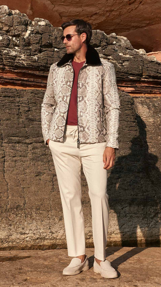 Man standing against a rocky wall wearing a patterned jacket and white pants.