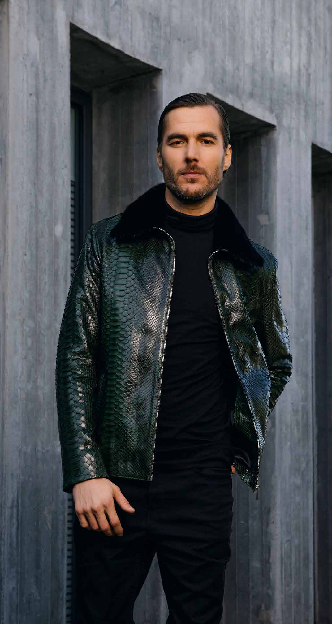 Man wearing a dark green leather jacket over a black shirt against a gray concrete wall.