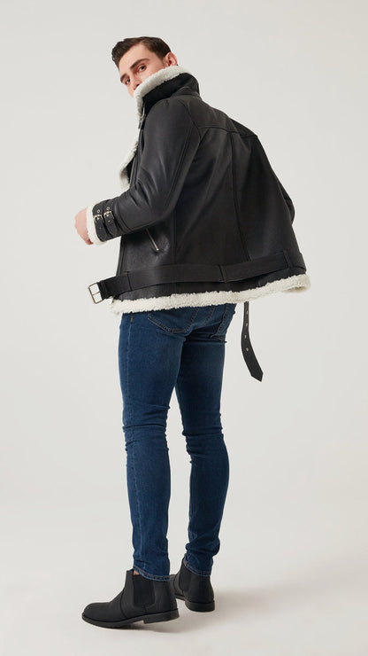 Man wearing a dark jacket with white fur lining, blue jeans, and black boots on a light gray background