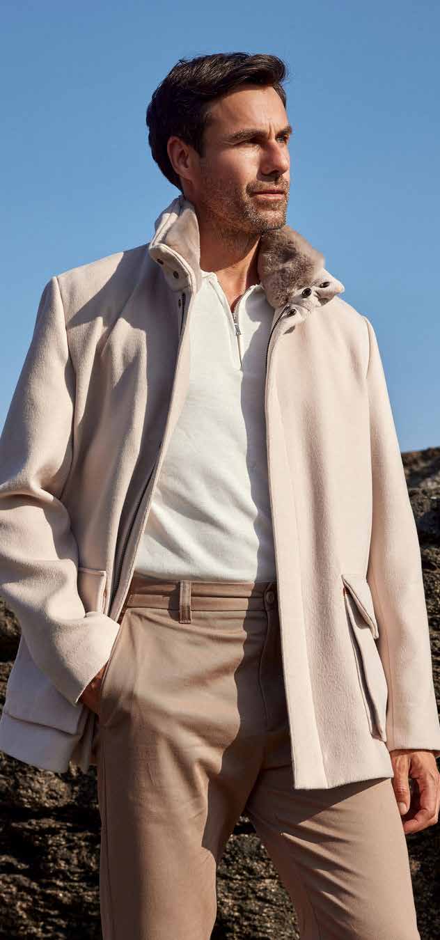 Man wearing a beige coat with fur collar and light brown pants against a clear blue sky.