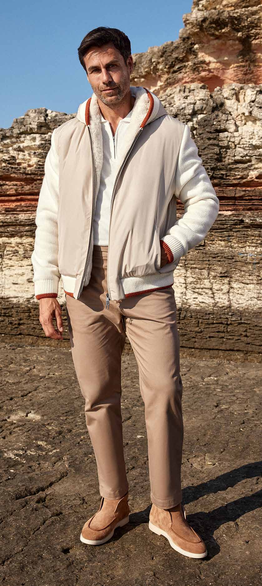 Man wearing a light gray jacket with a hood, beige pants, and brown shoes standing against a rocky cliff background.