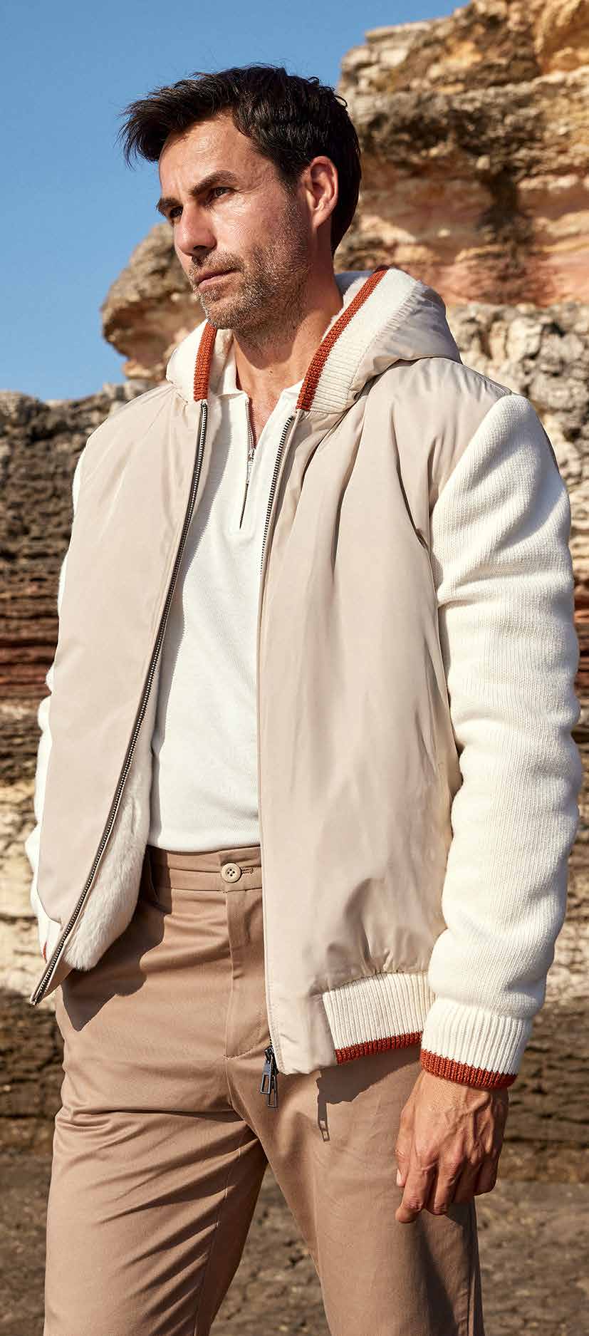 Man wearing a beige bomber jacket and brown pants standing in a rocky outdoor setting.