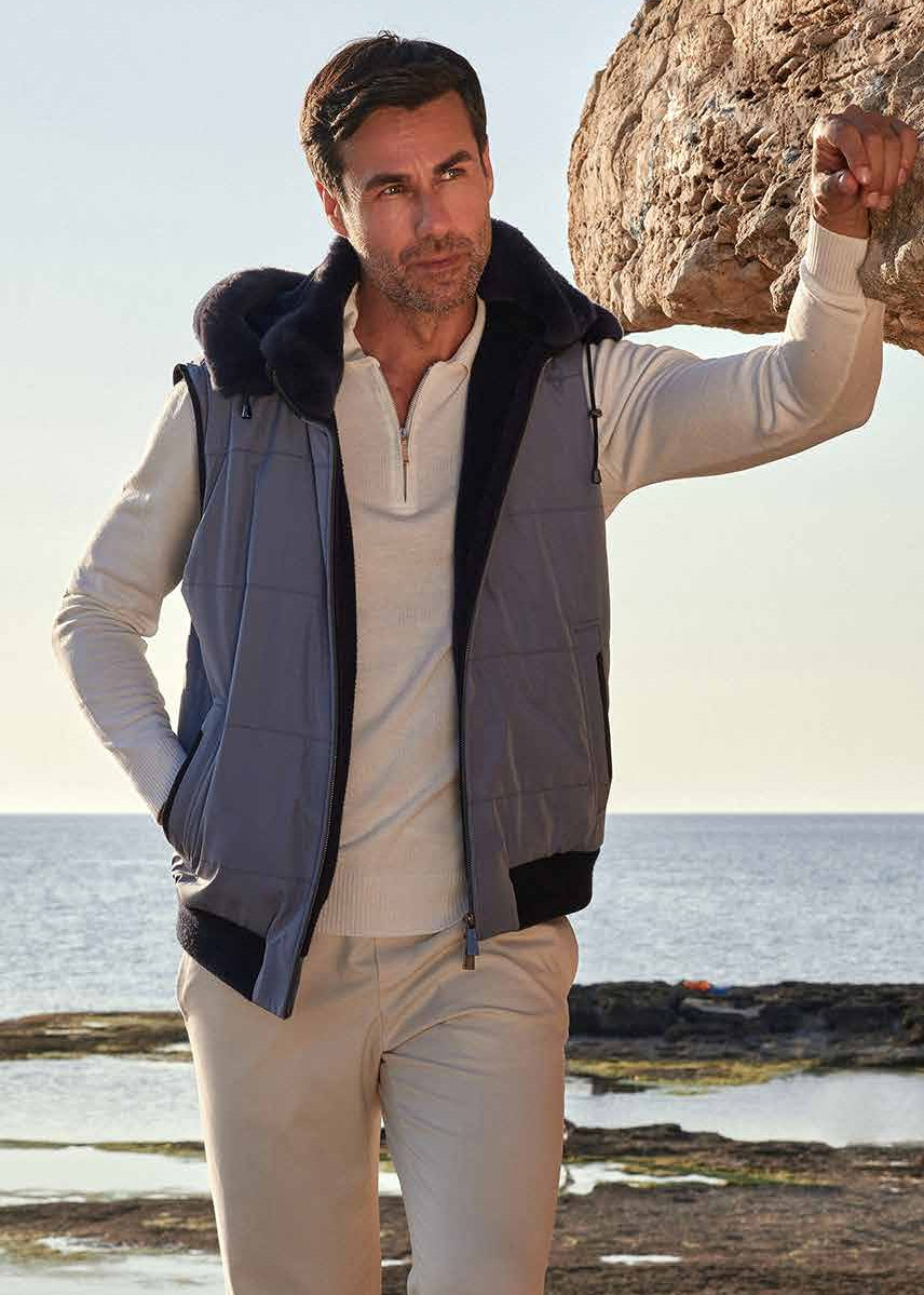 Man standing on a rocky beach wearing a light-colored outfit with a dark vest.