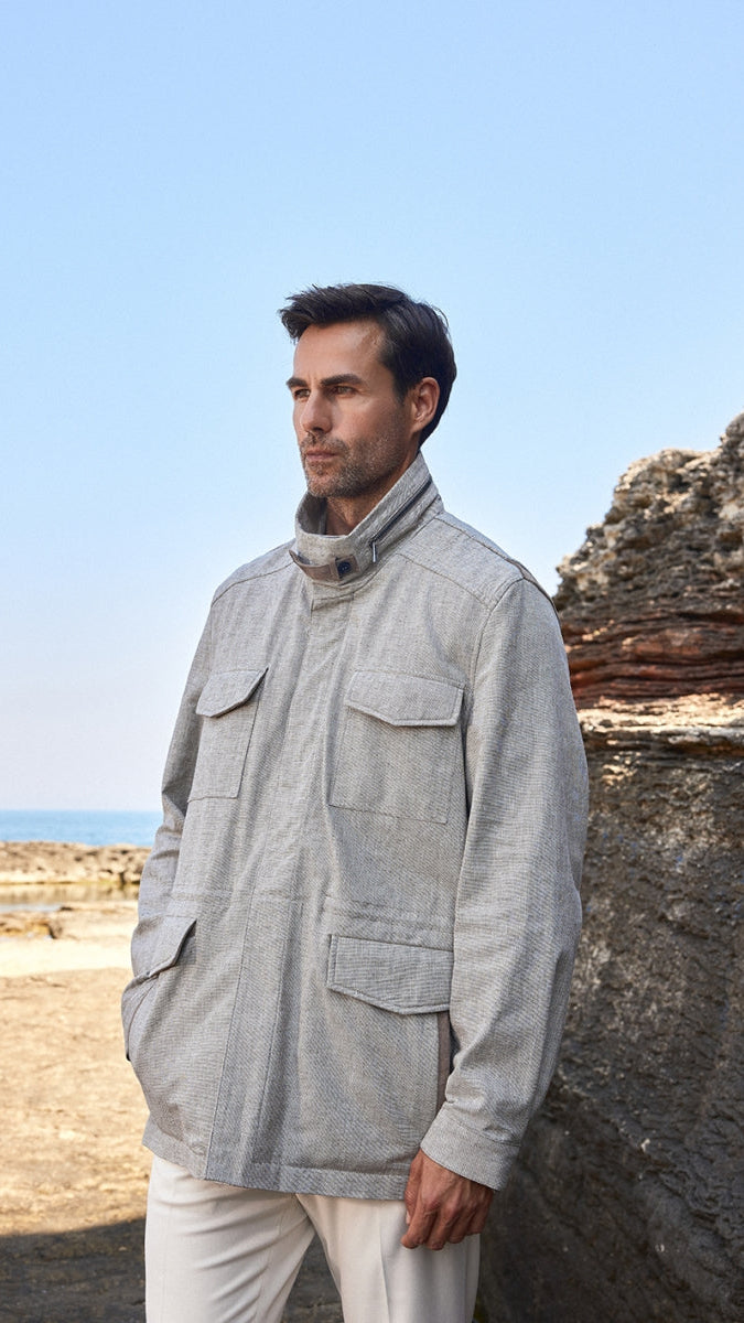 Man wearing a light gray jacket and white pants standing against a rocky coastal background.
