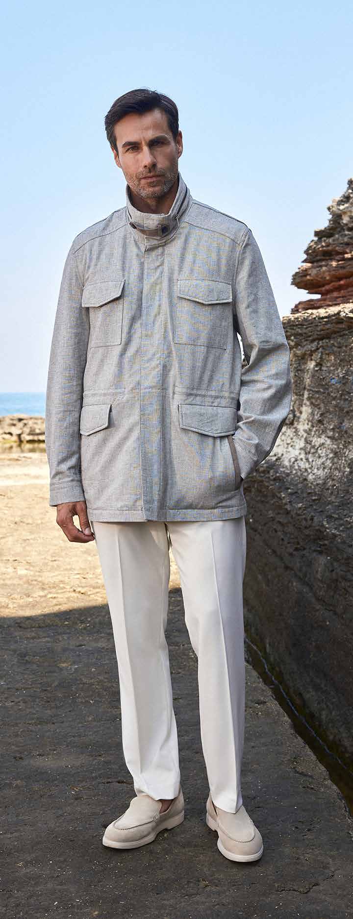 Man wearing a light gray jacket and white pants standing outdoors with a scenic background.