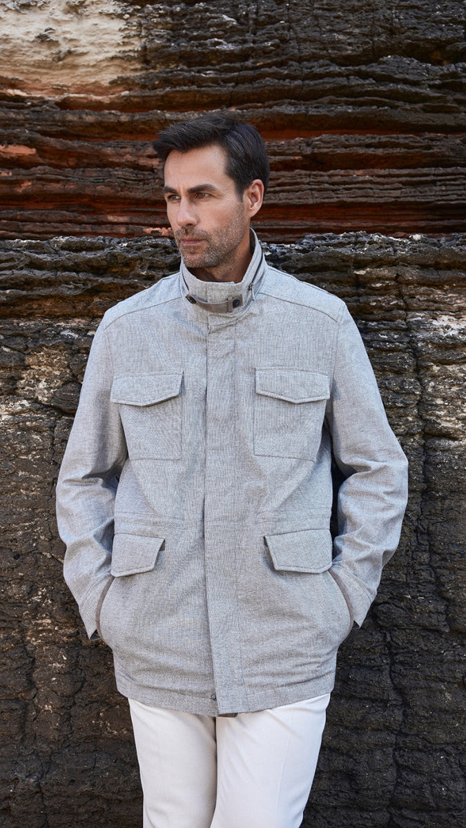 Man wearing a light gray jacket and white pants standing against a textured stone wall.