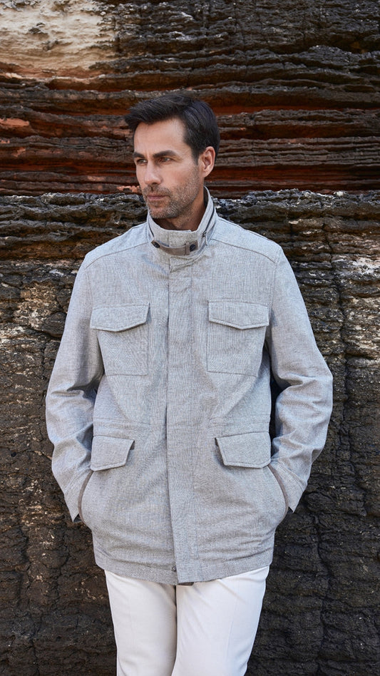 Man wearing a light gray jacket and white pants standing against a textured stone wall.